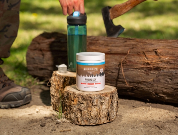 Photo of Morning Kick Container on a cut log with a green water bottle behind it. In the background is a person's hand on the water bottle and an ax stuck to the top on a long piece of wood.