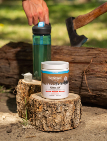 Photo of Morning Kick Container on a cut log with a green water bottle behind it. In the background is a person's hand on the water bottle and an ax stuck to the top on a long piece of wood.